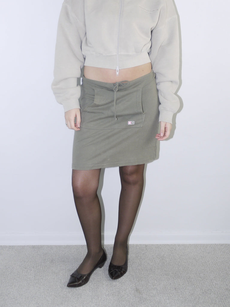 Vintage Y2K look featuring a cropped Alexander Wang zip-up fleece in beige, paired with an olive green mini skirt with front ruching and logo patch, styled with sheer tights and pointed flats.








