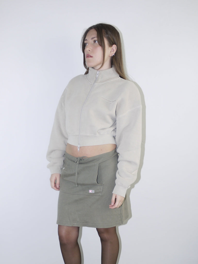 Vintage Y2K look featuring a cropped Alexander Wang zip-up fleece in beige, paired with an olive green mini skirt with front ruching and logo patch, styled with sheer tights and pointed flats.









