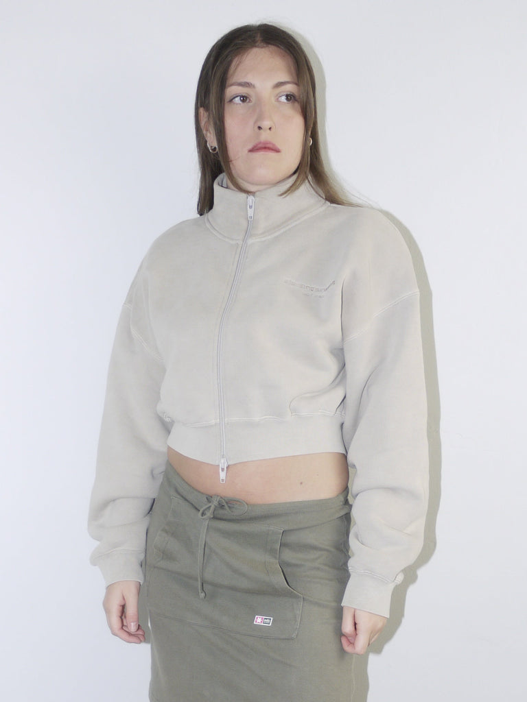 Vintage Y2K look featuring a cropped Alexander Wang zip-up fleece in beige, paired with an olive green mini skirt with front ruching and logo patch, styled with sheer tights and pointed flats.








