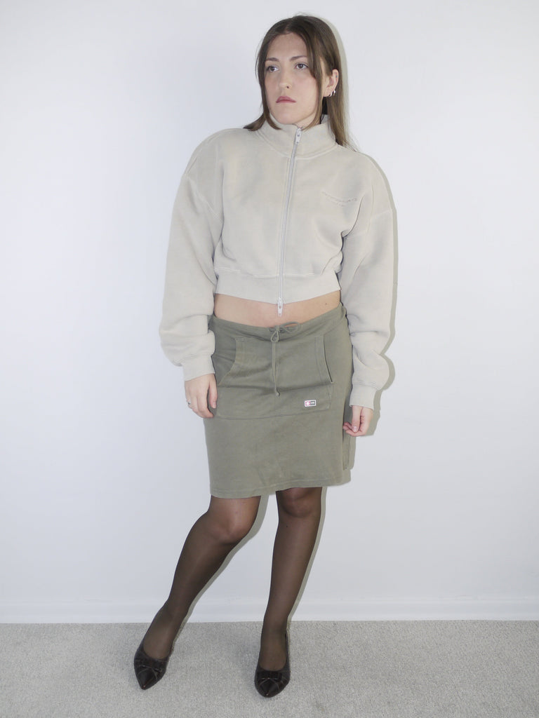 Vintage Y2K look featuring a cropped Alexander Wang zip-up fleece in beige, paired with an olive green mini skirt with front ruching and logo patch, styled with sheer tights and pointed flats.








