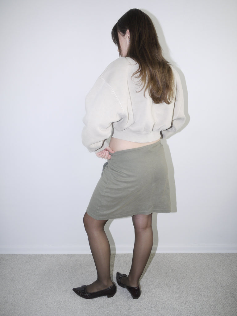 Vintage Y2K look featuring a cropped Alexander Wang zip-up fleece in beige, paired with an olive green mini skirt with front ruching and logo patch, styled with sheer tights and pointed flats.








