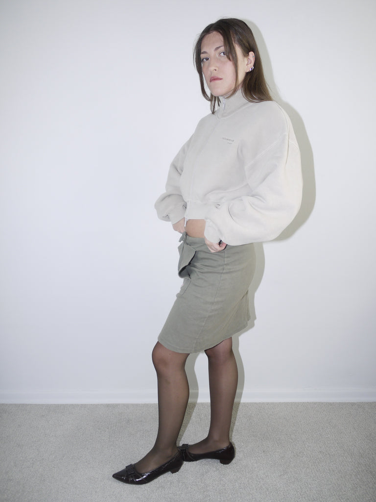Vintage Y2K look featuring a cropped Alexander Wang zip-up fleece in beige, paired with an olive green mini skirt with front ruching and logo patch, styled with sheer tights and pointed flats.








