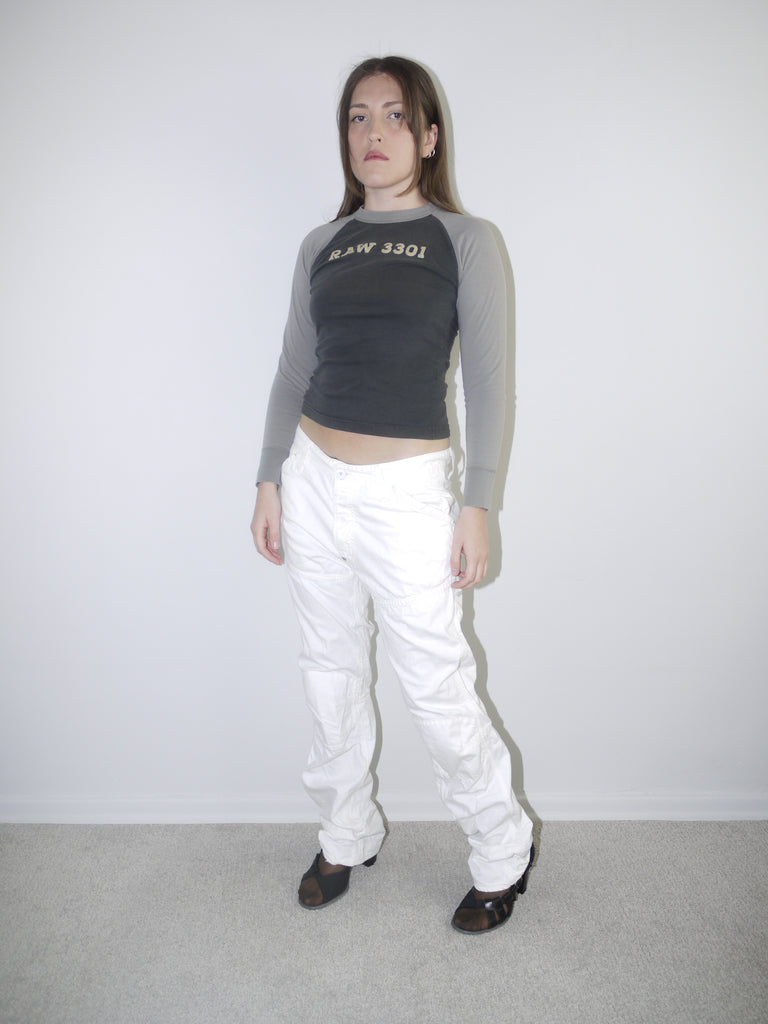 Back view of white low-rise cargo jeans with embroidered “96” detail on pocket, styled with black heeled boots and a cropped grey long sleeve top — bold Y2K revival look highlighting early 2000s denim silhouette, shot against white wall.

