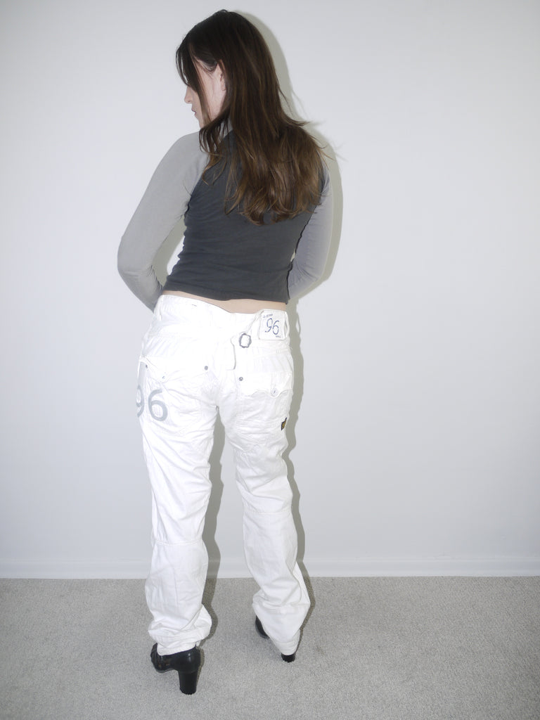Back view of white low-rise cargo jeans with embroidered “96” detail on pocket, styled with black heeled boots and a cropped grey long sleeve top — bold Y2K revival look highlighting early 2000s denim silhouette, shot against white wall.

