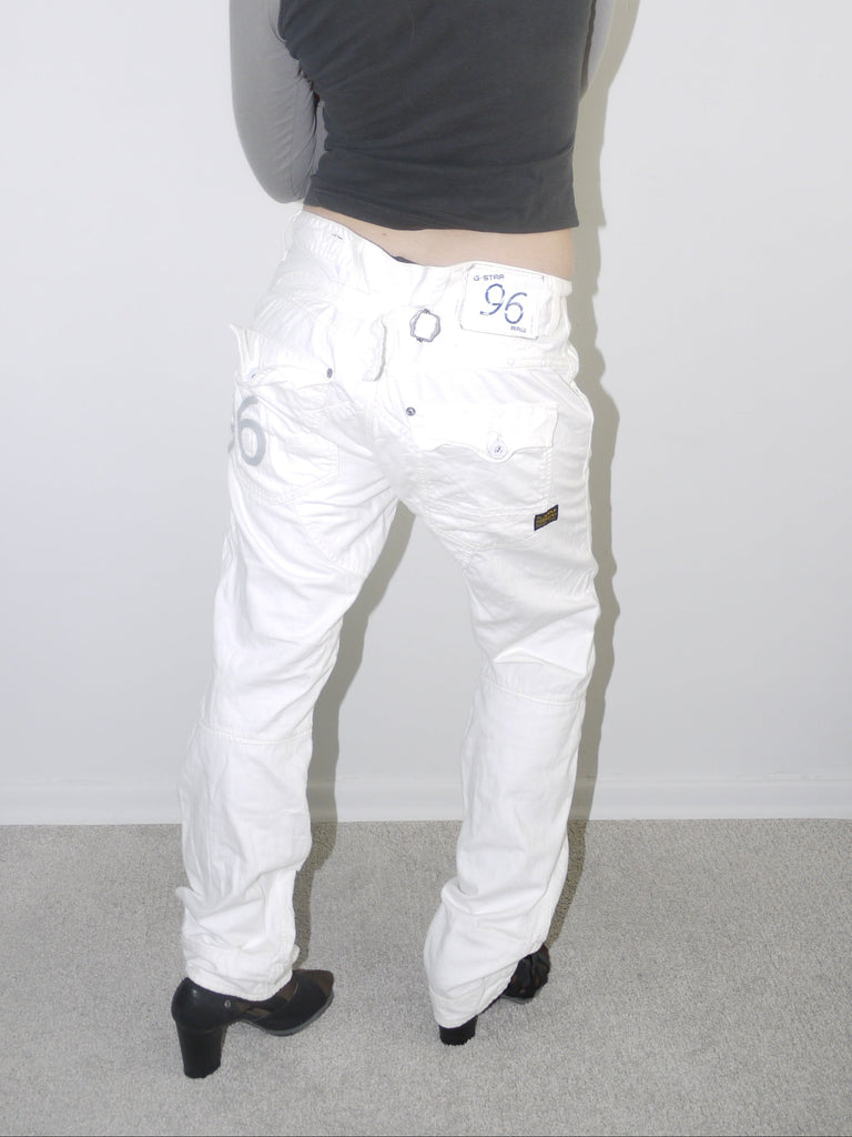 Back view of white low-rise cargo jeans with embroidered “96” detail on pocket, styled with black heeled boots and a cropped grey long sleeve top — bold Y2K revival look highlighting early 2000s denim silhouette, shot against white wall.

