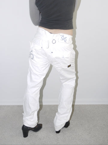 Back view of white low-rise cargo jeans with embroidered “96” detail on pocket, styled with black heeled boots and a cropped grey long sleeve top — bold Y2K revival look highlighting early 2000s denim silhouette, shot against white wall.

