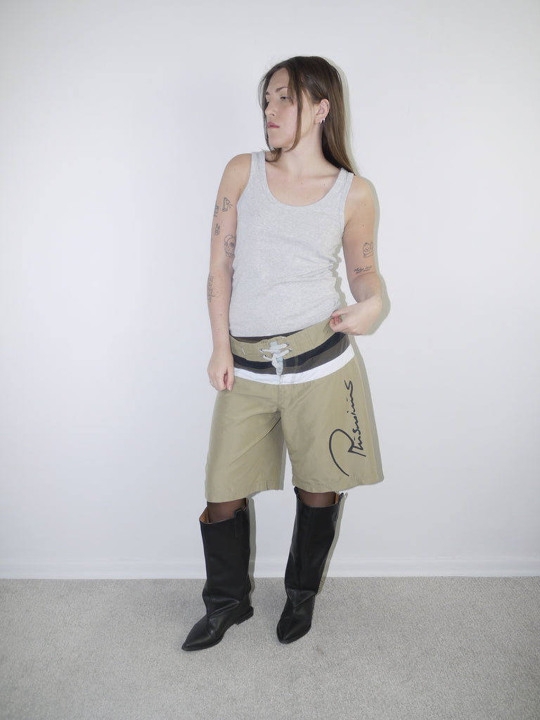Model wearing beige Plus Minus logo tech shorts with bold contrast waistband and drawstring closure, styled with a fitted grey ribbed tank top and black knee-high leather boots — Y2K streetwear look with utility and branding focus, photographed against white background.