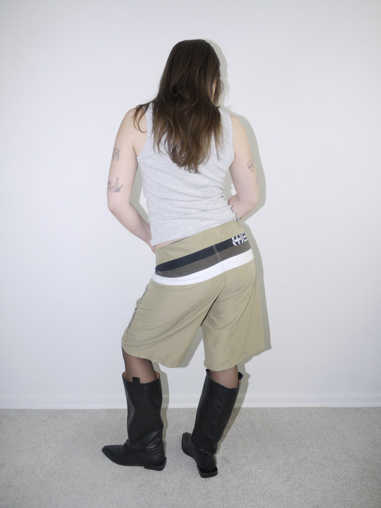 Model wearing beige Plus Minus logo tech shorts with bold contrast waistband and drawstring closure, styled with a fitted grey ribbed tank top and black knee-high leather boots — Y2K streetwear look with utility and branding focus, photographed against white background.