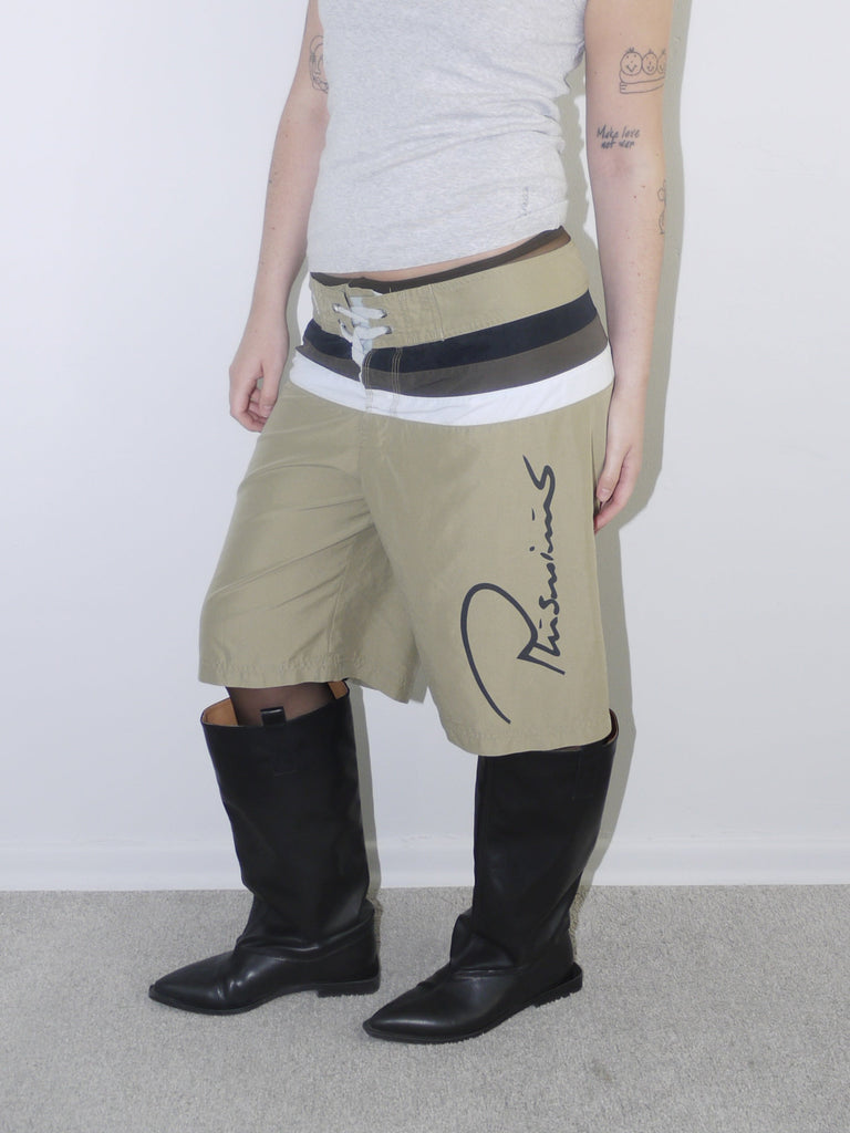Model wearing beige Plus Minus logo tech shorts with bold contrast waistband and drawstring closure, styled with a fitted grey ribbed tank top and black knee-high leather boots — Y2K streetwear look with utility and branding focus, photographed against white background.