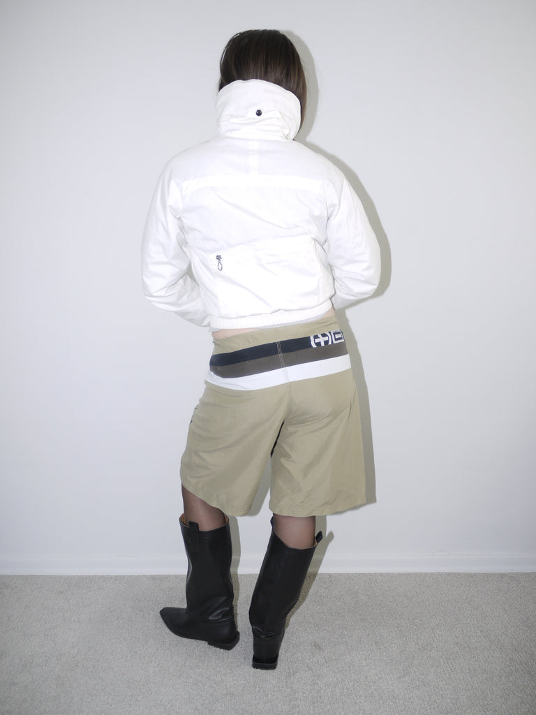 Model wearing a white cropped zip-up windbreaker jacket paired with beige tech-fabric shorts featuring graphic details, wide contrast waistband and black leather knee-high boots — bold futuristic streetwear look with Y2K and cyber influences, photographed against plain background.