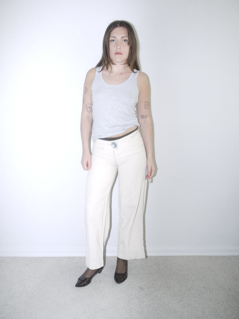 Model wearing cream wide-leg Cop Copine trousers with mid-rise waist, styled with black tights, pointed flats and a fitted grey ribbed tank top — soft minimal Y2K-inspired look with subtle Parisian edge, photographed against white background.