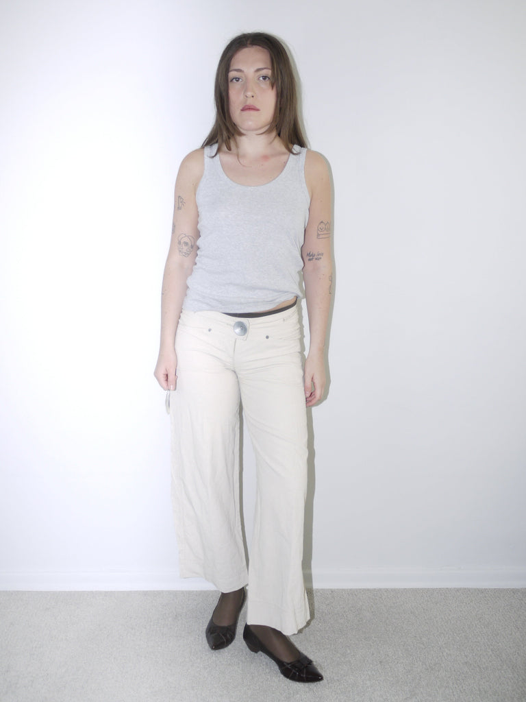 Model wearing cream wide-leg Cop Copine trousers with mid-rise waist, styled with black tights, pointed flats and a fitted grey ribbed tank top — soft minimal Y2K-inspired look with subtle Parisian edge, photographed against white background.