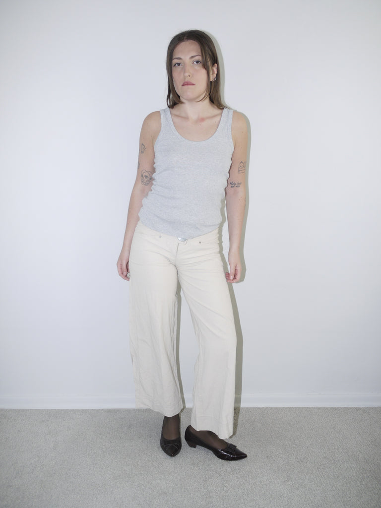 Model wearing cream wide-leg Cop Copine trousers with mid-rise waist, styled with black tights, pointed flats and a fitted grey ribbed tank top — soft minimal Y2K-inspired look with subtle Parisian edge, photographed against white background.