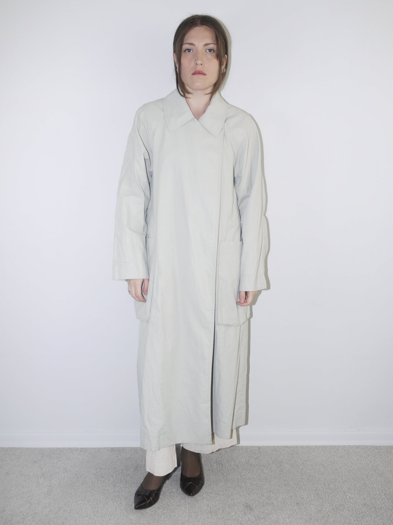 Model wearing a long light grey COS coat with minimal collar design, oversized fit and hidden closure, styled over wide-leg cream trousers and black pointed flats — clean Scandinavian-inspired silhouette with architectural tailoring, photographed against white wall.