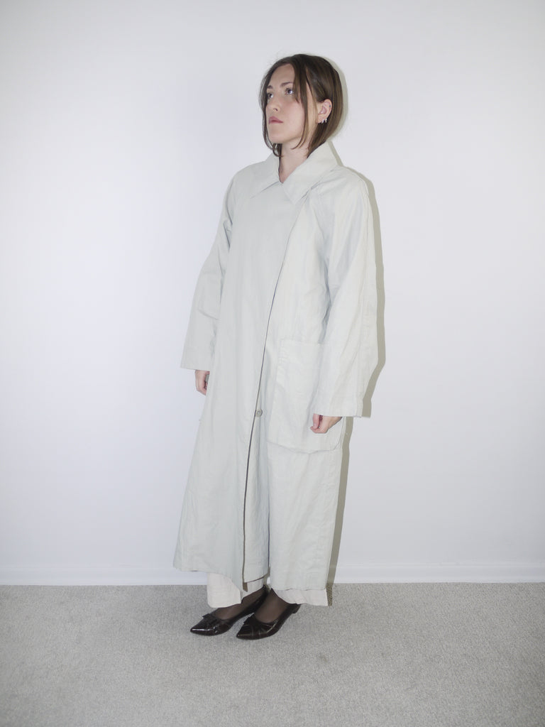 Model wearing a long light grey COS coat with minimal collar design, oversized fit and hidden closure, styled over wide-leg cream trousers and black pointed flats — clean Scandinavian-inspired silhouette with architectural tailoring, photographed against white wall.