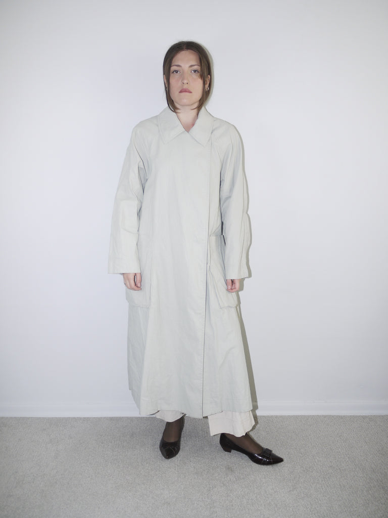 Model wearing a long light grey COS coat with minimal collar design, oversized fit and hidden closure, styled over wide-leg cream trousers and black pointed flats — clean Scandinavian-inspired silhouette with architectural tailoring, photographed against white wall.