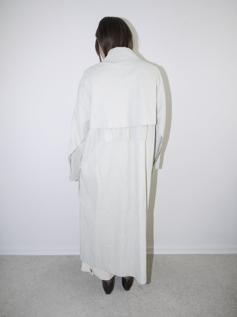 Model wearing a long light grey COS coat with minimal collar design, oversized fit and hidden closure, styled over wide-leg cream trousers and black pointed flats — clean Scandinavian-inspired silhouette with architectural tailoring, photographed against white wall.