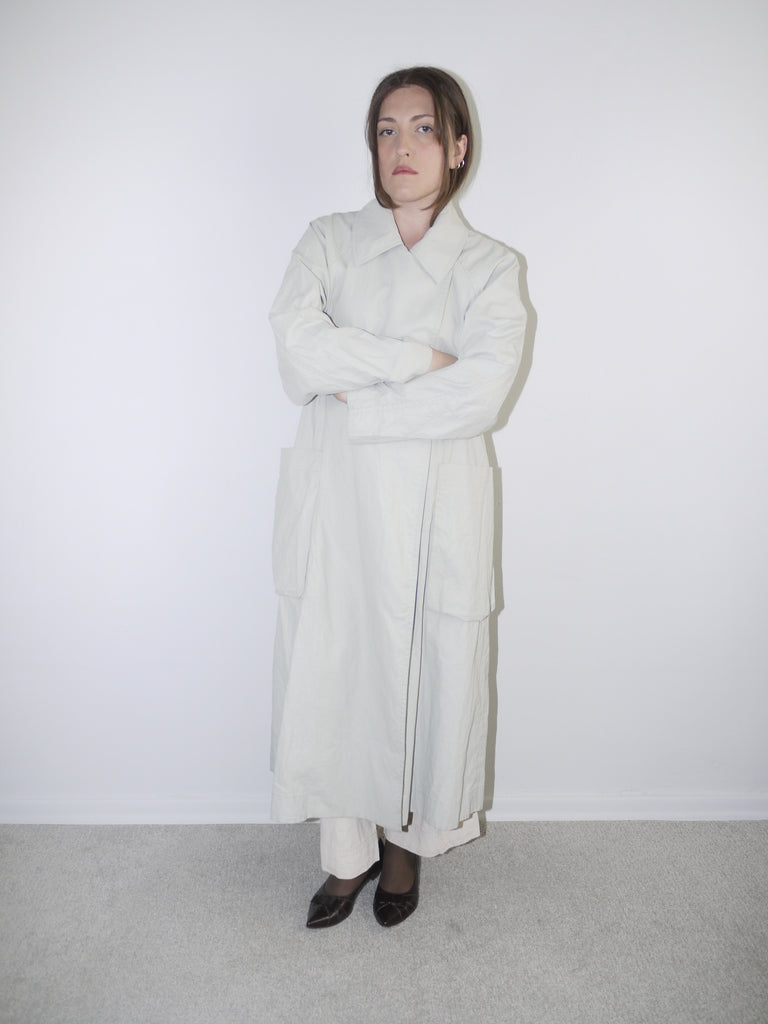 Model wearing a long light grey COS coat with minimal collar design, oversized fit and hidden closure, styled over wide-leg cream trousers and black pointed flats — clean Scandinavian-inspired silhouette with architectural tailoring, photographed against white wall.