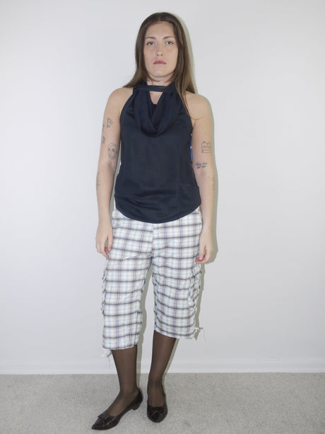 Model wearing a sleeveless navy draped-neck top paired with white and navy plaid knee-length drawstring shorts, styled with sheer black tights and pointed patent flats — experimental layered look with Y2K influence, photographed against plain white background.
