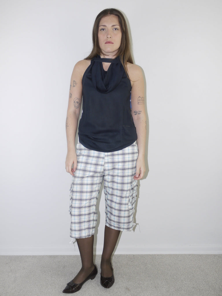 Model wearing a sleeveless navy draped-neck top paired with white and navy plaid knee-length drawstring shorts, styled with sheer black tights and pointed patent flats — experimental layered look with Y2K influence, photographed against plain white background.