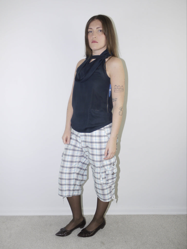 Model wearing a sleeveless navy draped-neck top paired with white and navy plaid knee-length drawstring shorts, styled with sheer black tights and pointed patent flats — experimental layered look with Y2K influence, photographed against plain white background.