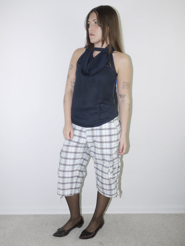 Model wearing a sleeveless navy draped-neck top paired with white and navy plaid knee-length drawstring shorts, styled with sheer black tights and pointed patent flats — experimental layered look with Y2K influence, photographed against plain white background.
