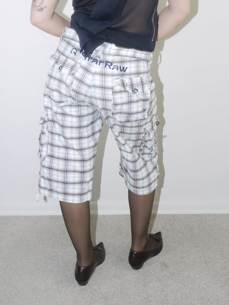 Model wearing a sleeveless navy draped-neck top paired with white and navy plaid knee-length drawstring shorts, styled with sheer black tights and pointed patent flats — experimental layered look with Y2K influence, photographed against plain white background.