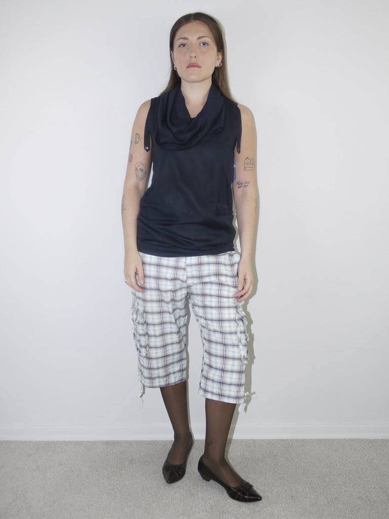Model wearing a sleeveless navy draped-neck top paired with white and navy plaid knee-length drawstring shorts, styled with sheer black tights and pointed patent flats — experimental layered look with Y2K influence, photographed against plain white background.