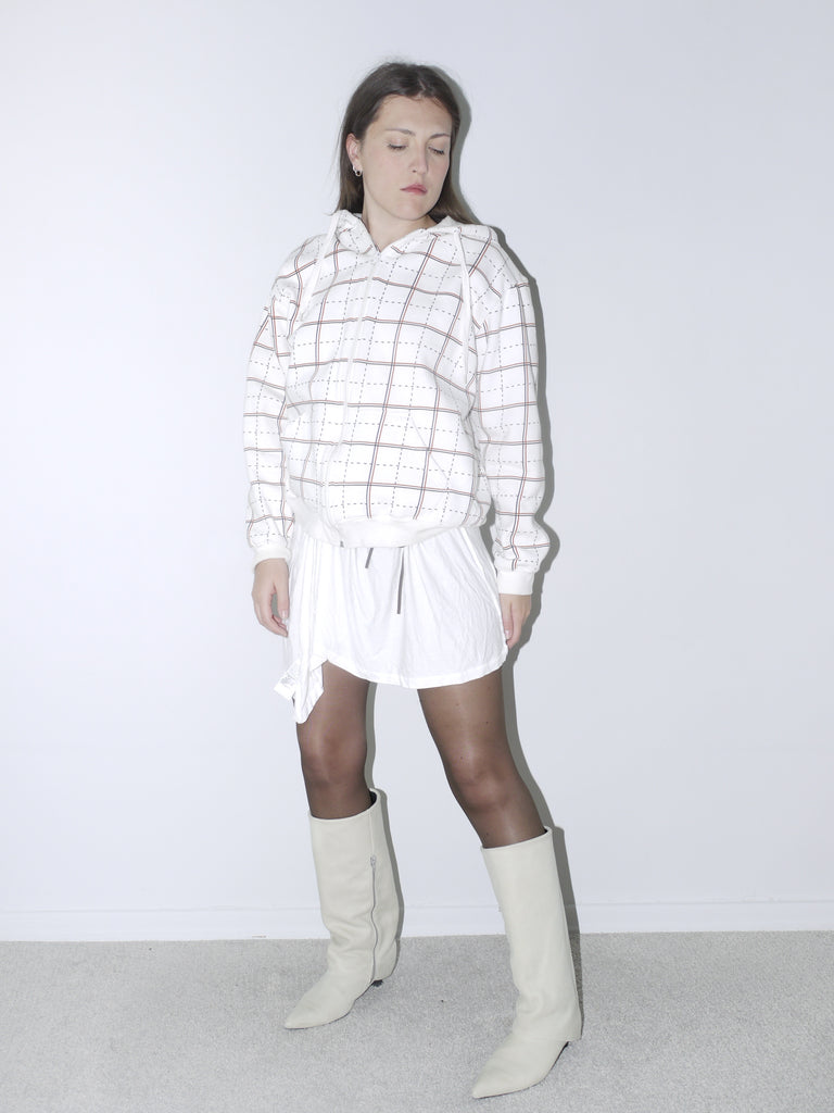 Model wearing a white grid-pattern hoodie paired with a white drawstring mini skirt, sheer black tights, and off-white pointed knee-high boots. A clean, contemporary outfit with retro sportswear vibes, styled against a plain white backdrop.
