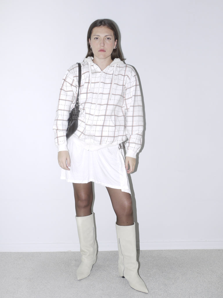 Model wearing a white grid-pattern hoodie paired with a white drawstring mini skirt, sheer black tights, and off-white pointed knee-high boots. A clean, contemporary outfit with retro sportswear vibes, styled against a plain white backdrop.