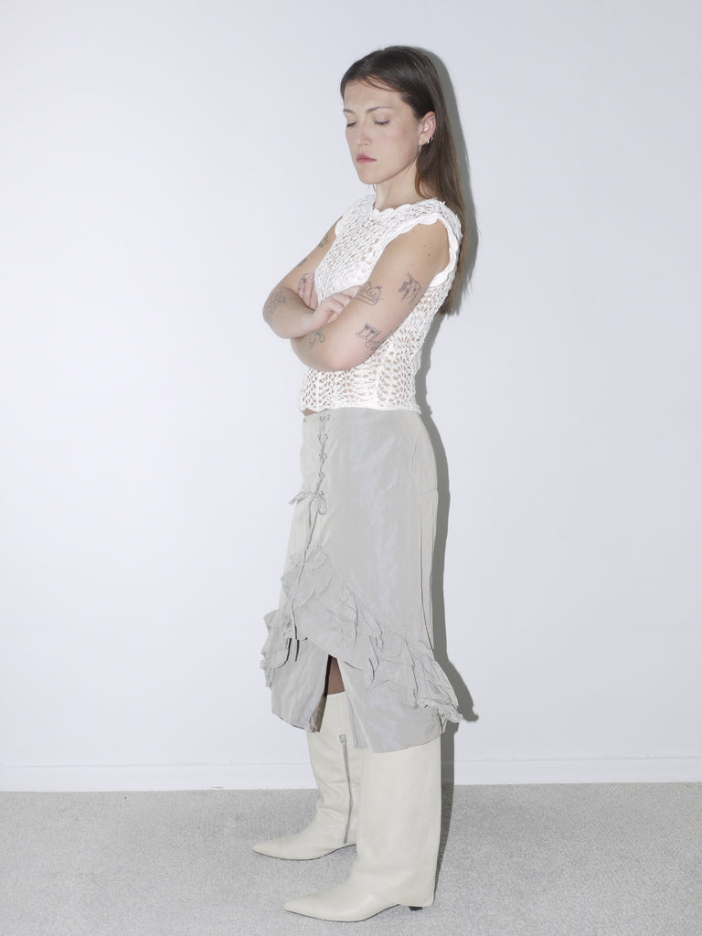 Delicate white crochet top paired with a layered, ruffled midi skirt in muted taupe, styled with white pointed knee-high boots for a soft yet structured vintage-inspired look