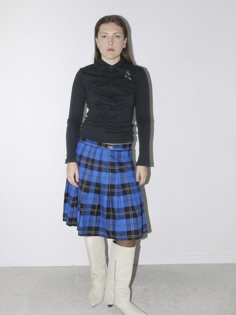 Black fitted long-sleeve top with embroidered collar details, paired with a bold blue and black plaid pleated skirt and white pointed knee-high boots for a preppy vintage-inspired ensemble.