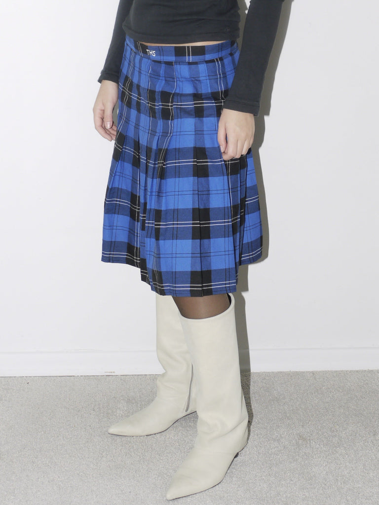 Black fitted long-sleeve top with embroidered collar details, paired with a bold blue and black plaid pleated skirt and white pointed knee-high boots for a preppy vintage-inspired ensemble.