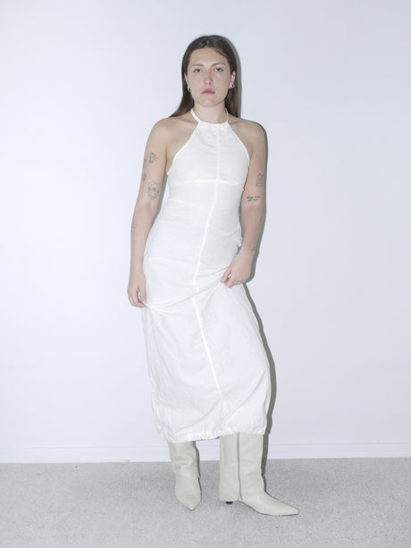 Model wearing a white halter-neck Diesel dress with exposed seam detailing and a softly structured silhouette, paired with cream-colored pointed cowboy boots. The look channels early-2000s minimalism with a touch of western edge. Shot against a plain white wall.