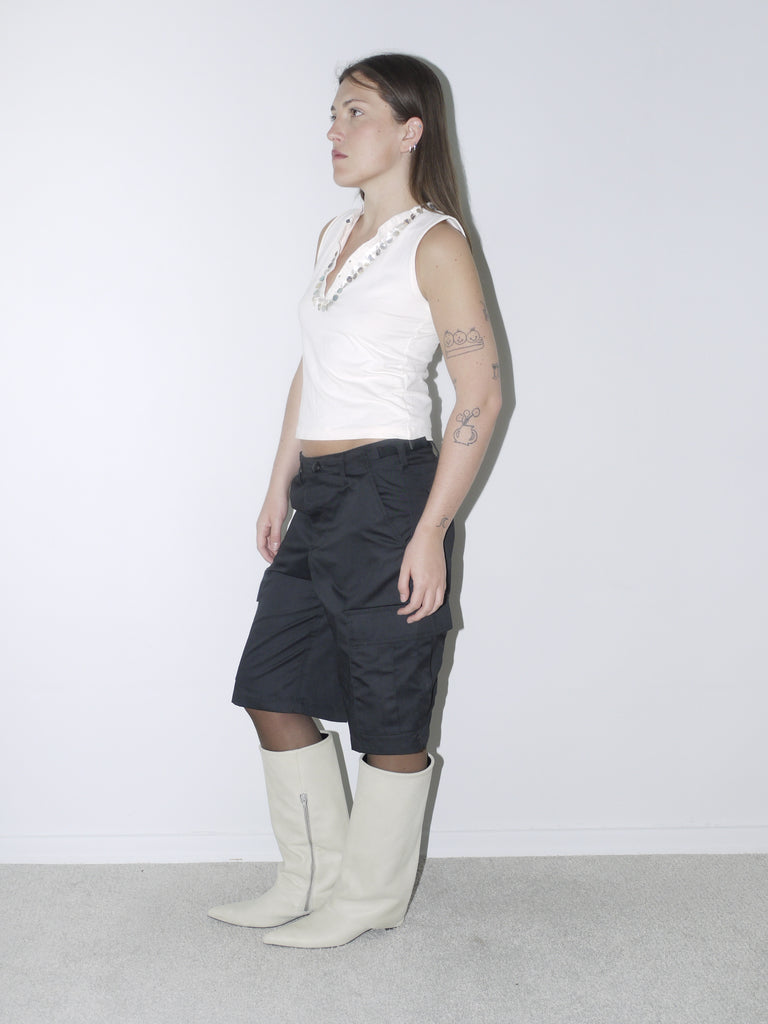 Sleeveless ivory top with pearl embellishment detail, paired with black satin bermuda cargo shorts and cream pointed boots – a unique mix of elegance and utilitarian edge.