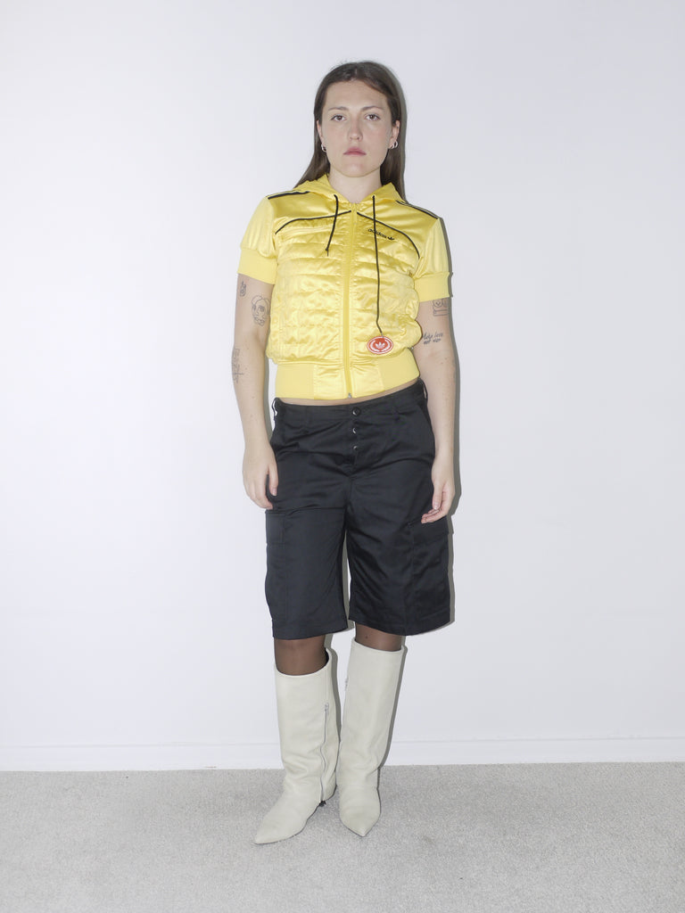 Bold vintage look featuring a cropped shiny yellow Adidas Originals zip hoodie with short sleeves, paired with black satin bermuda cargo shorts and off-white pointed boots – playful and sporty with Y2K energy.