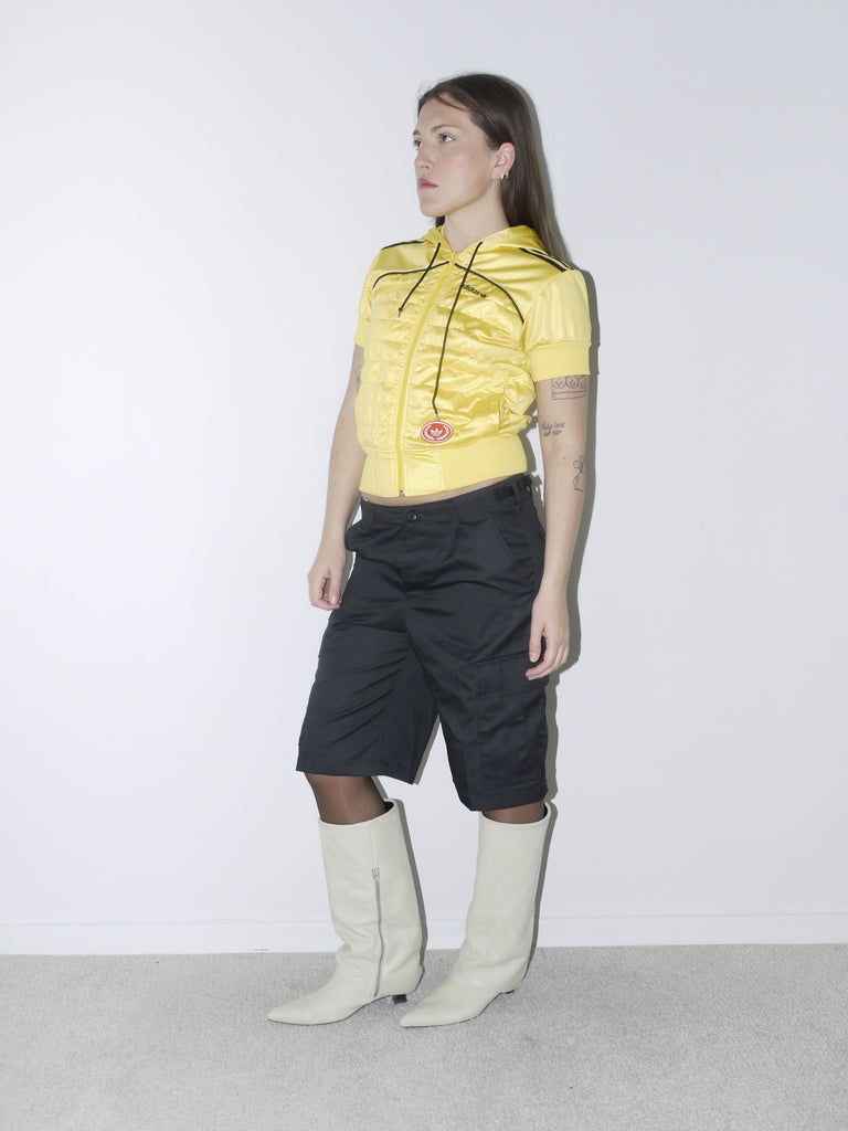 Bold vintage look featuring a cropped shiny yellow Adidas Originals zip hoodie with short sleeves, paired with black satin bermuda cargo shorts and off-white pointed boots – playful and sporty with Y2K energy.