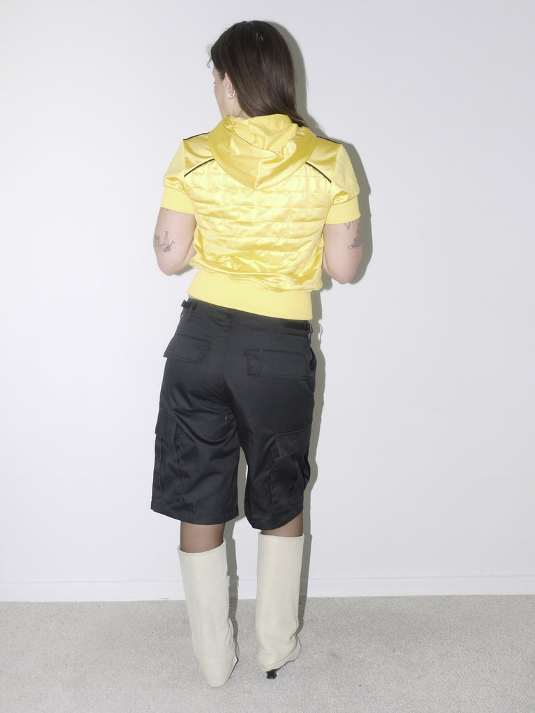Bold vintage look featuring a cropped shiny yellow Adidas Originals zip hoodie with short sleeves, paired with black satin bermuda cargo shorts and off-white pointed boots – playful and sporty with Y2K energy.
