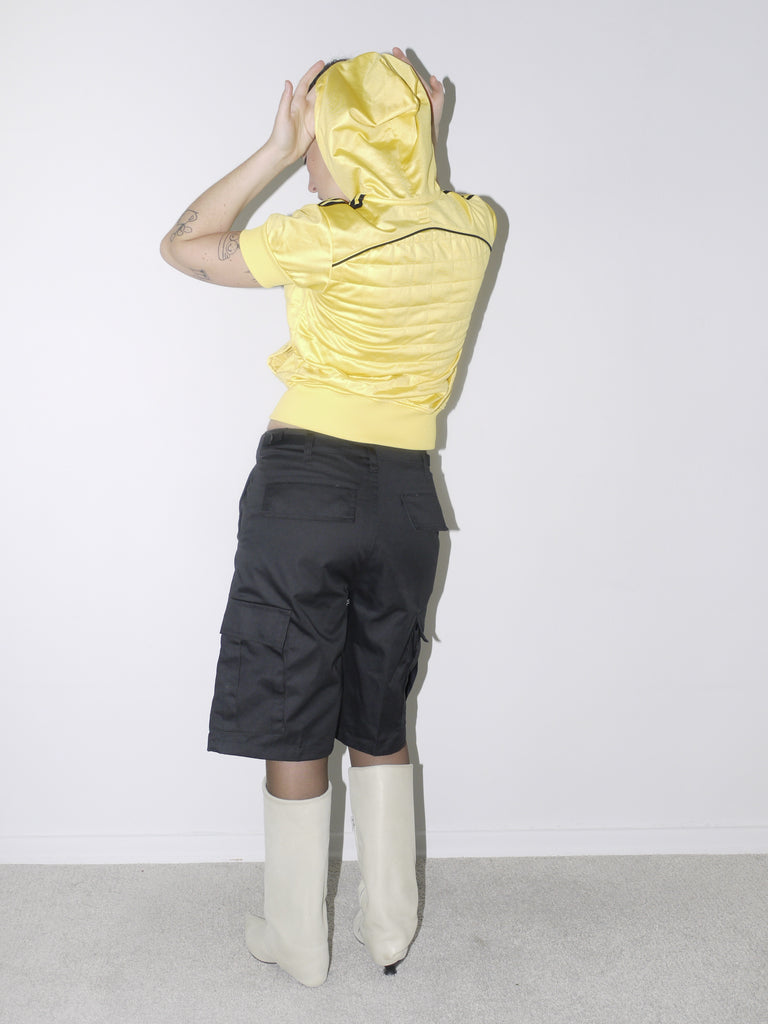Bold vintage look featuring a cropped shiny yellow Adidas Originals zip hoodie with short sleeves, paired with black satin bermuda cargo shorts and off-white pointed boots – playful and sporty with Y2K energy.