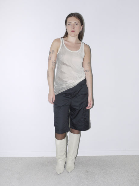 Minimal Y2K-inspired outfit featuring a sheer ivory racerback tank top with abstract print details, paired with black satin cargo-style bermuda shorts and off-white pointed boots – clean, edgy, and effortlessly cool.