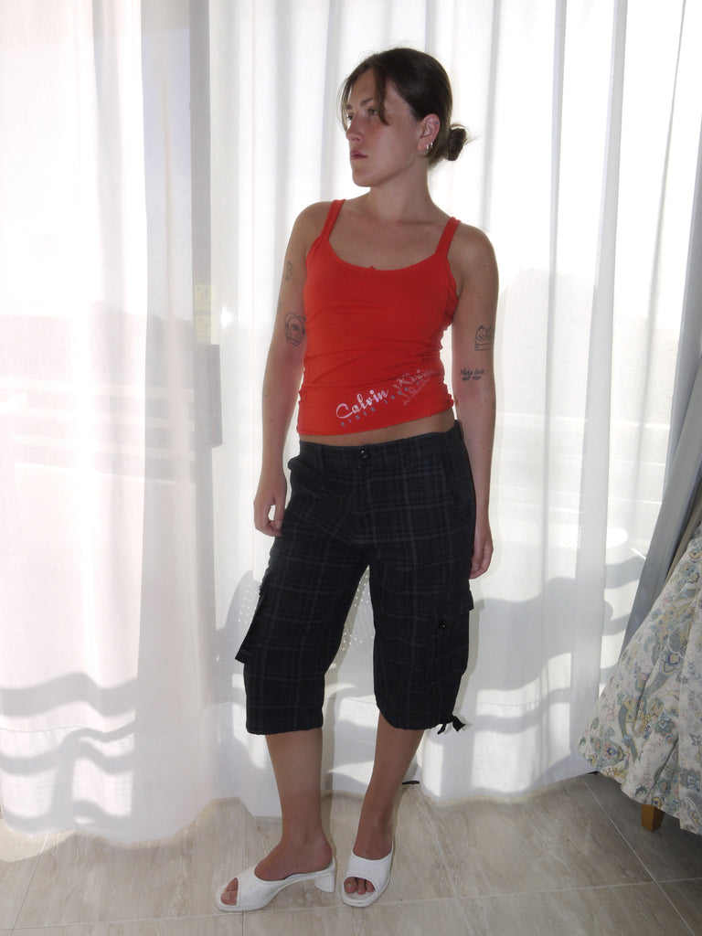 Model wearing a fitted red tank top with subtle graphic detailing, paired with low-rise dark capri pants featuring a checkered pattern and drawstring hems. Styled with black-and-white printed slingback heels, creating a sharp Y2K-inspired contrast. Shot against a plain white wall.