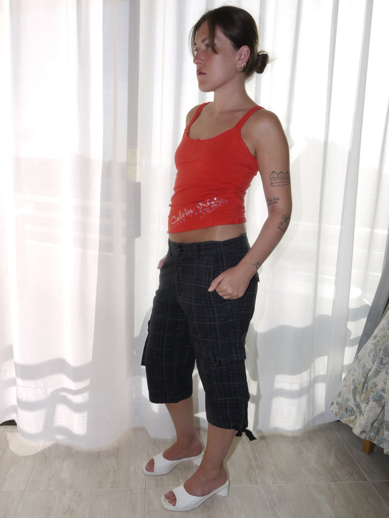Model wearing a fitted red tank top with subtle graphic detailing, paired with low-rise dark capri pants featuring a checkered pattern and drawstring hems. Styled with black-and-white printed slingback heels, creating a sharp Y2K-inspired contrast. Shot against a plain white wall.