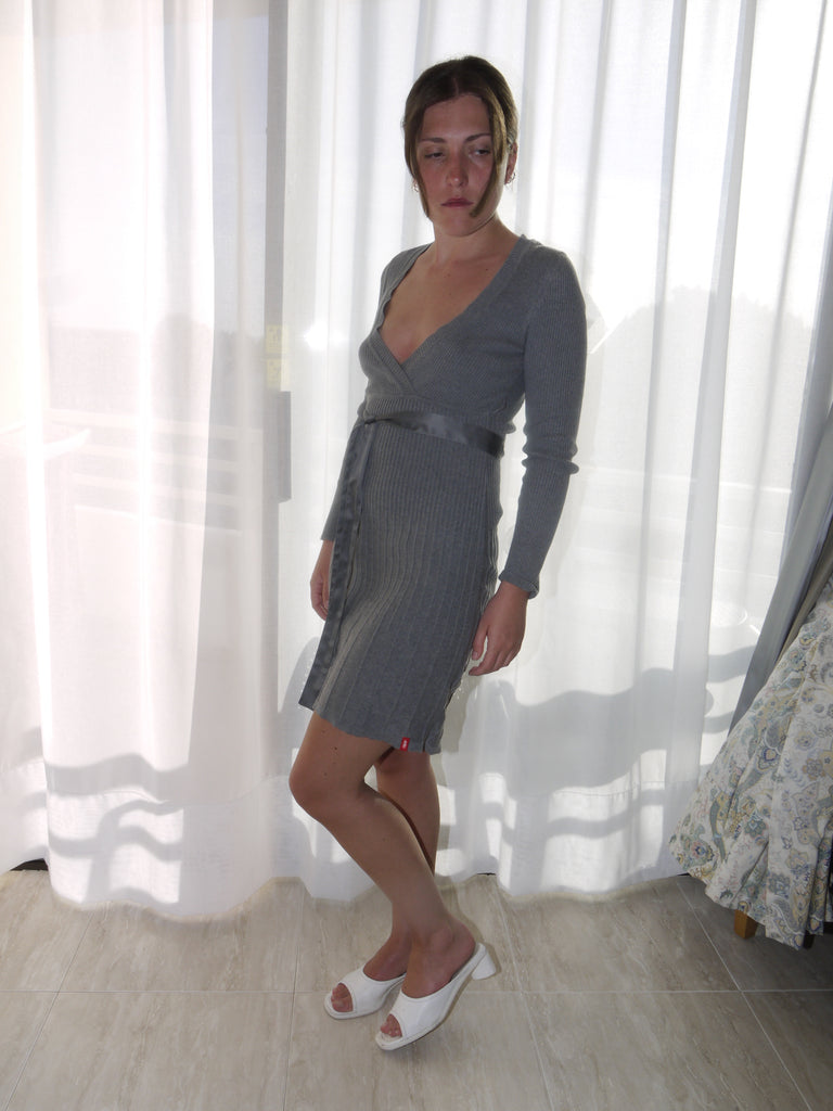 Model wearing a fitted grey ribbed knit dress with long sleeves, a deep V-neckline, and a tie-belt at the waist. Styled with tan ankle boots for a warm, casual finish. Shot against a plain white wall.