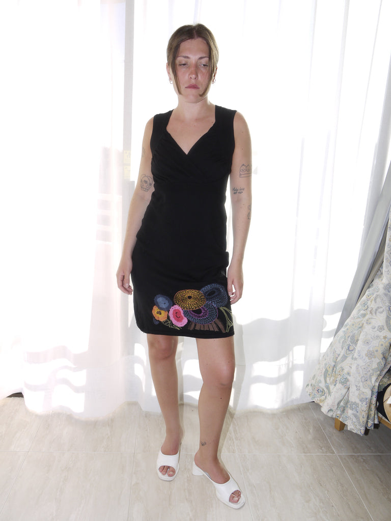 Model wearing a sleeveless black Desigual dress with a deep V-neckline and colorful floral embroidery along the hem. Styled with tan ankle boots for a bold contrast between playful detailing and minimal silhouette. Shot against a plain white background