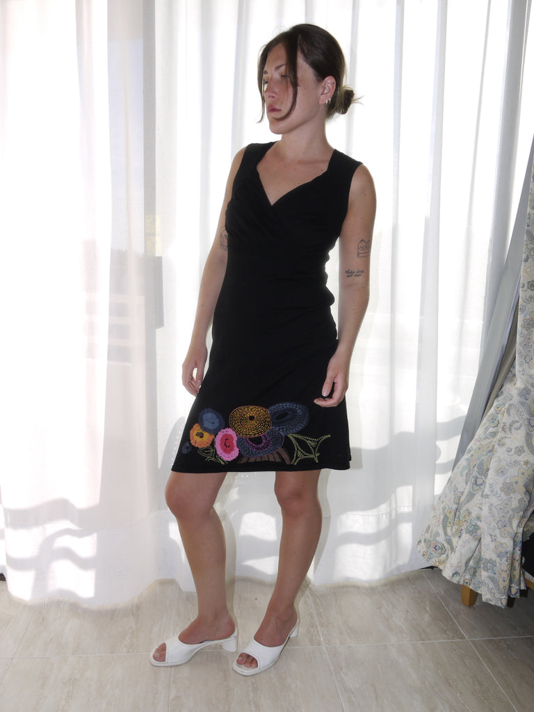 Model wearing a sleeveless black Desigual dress with a deep V-neckline and colorful floral embroidery along the hem. Styled with tan ankle boots for a bold contrast between playful detailing and minimal silhouette. Shot against a plain white background