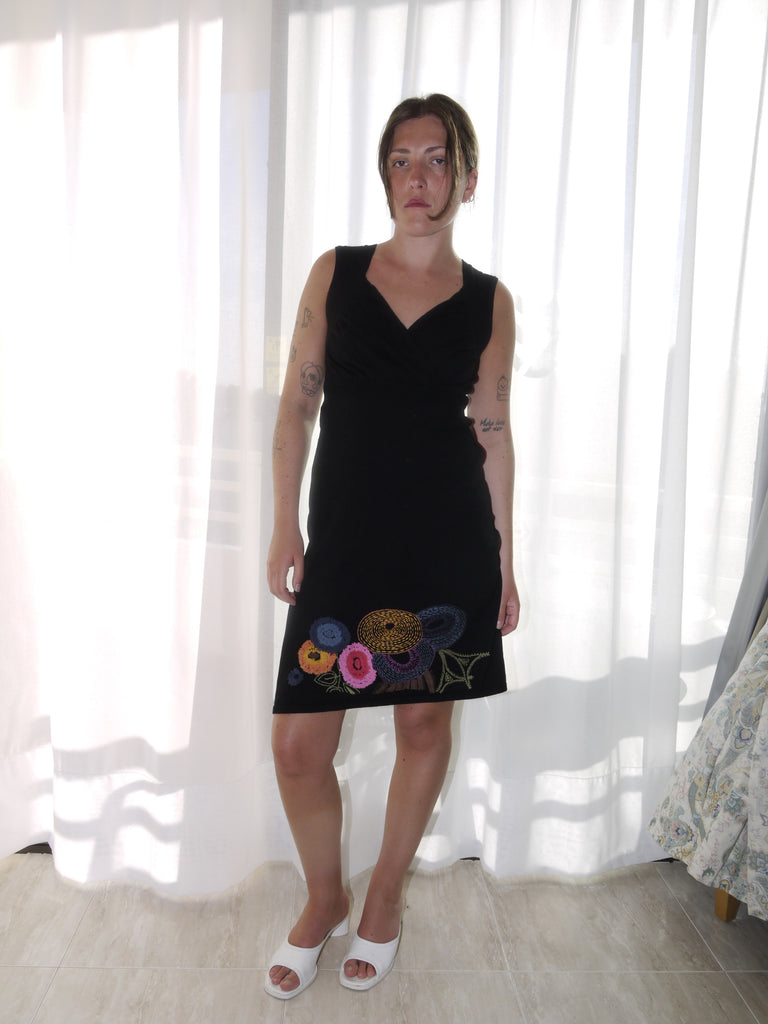 Model wearing a sleeveless black Desigual dress with a deep V-neckline and colorful floral embroidery along the hem. Styled with tan ankle boots for a bold contrast between playful detailing and minimal silhouette. Shot against a plain white background