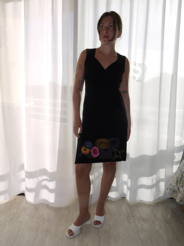 Model wearing a sleeveless black Desigual dress with a deep V-neckline and colorful floral embroidery along the hem. Styled with tan ankle boots for a bold contrast between playful detailing and minimal silhouette. Shot against a plain white background