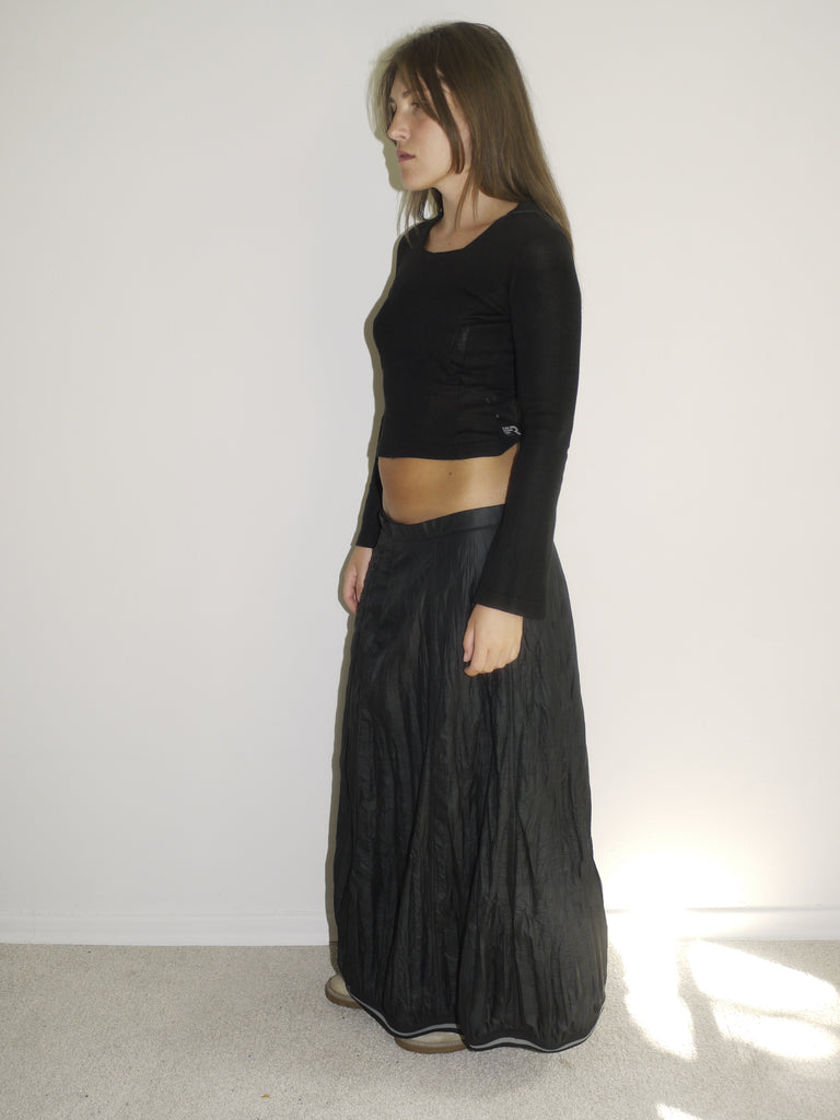 Model wearing a fitted black hooded top with long sleeves and a cropped silhouette, paired with a flowy, slightly crinkled black maxi skirt. Styled with beige slip-on shoes, the look blends gothic minimalism with early-2000s casual drama. Shot from the side against a plain white wall with natural light.