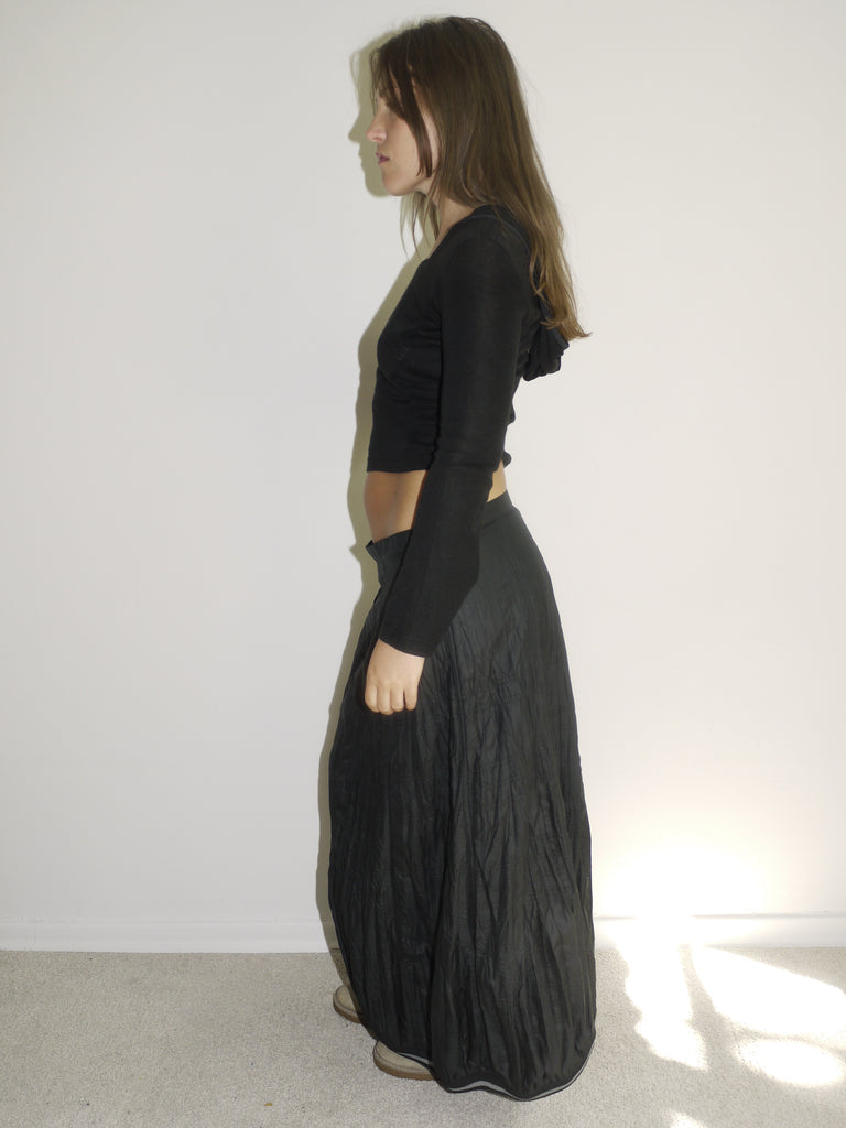 Model wearing a fitted black hooded top with long sleeves and a cropped silhouette, paired with a flowy, slightly crinkled black maxi skirt. Styled with beige slip-on shoes, the look blends gothic minimalism with early-2000s casual drama. Shot from the side against a plain white wall with natural light.