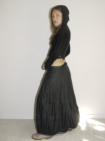 Model wearing a fitted black hooded top with long sleeves and a cropped silhouette, paired with a flowy, slightly crinkled black maxi skirt. Styled with beige slip-on shoes, the look blends gothic minimalism with early-2000s casual drama. Shot from the side against a plain white wall with natural light.